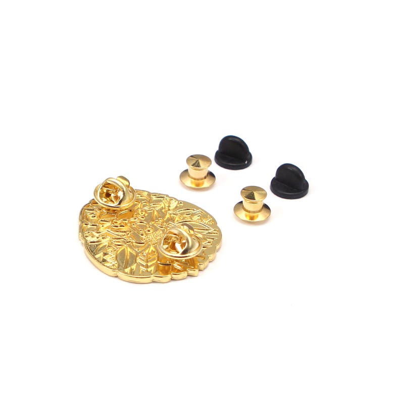 Puede incluir: Un pin de metal dorado con un intrincado dise&ntilde;o floral. El pin tiene dos respaldos de alfiler adjuntos, junto con tres respaldos de alfiler dorados adicionales y tres cierres de goma negros. El pin est&aacute; sobre un fondo blanco.