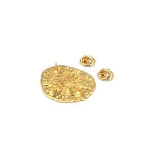 Puede incluir: Un pin de metal dorado con un intrincado dise&ntilde;o floral. El pin tiene un solo poste y dos cierres de pin adicionales. El pin est&aacute; sobre un fondo blanco.
