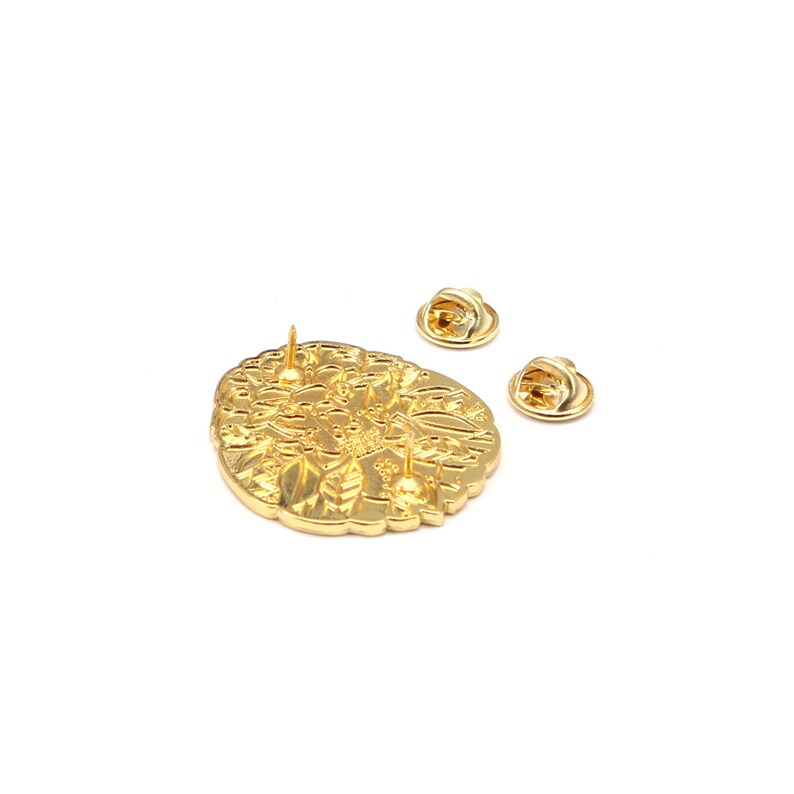 Puede incluir: Un pin de metal dorado con un intrincado dise&ntilde;o floral. El pin tiene un solo poste y dos cierres de pin adicionales. El pin est&aacute; sobre un fondo blanco.