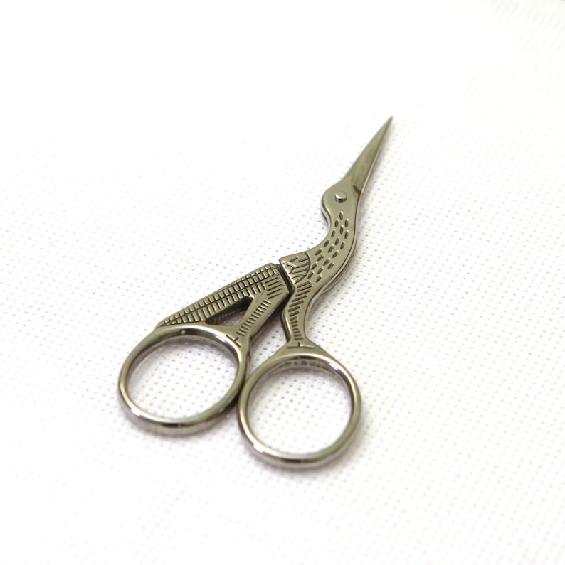 May include: A pair of silver-colored stork scissors. The scissors have a detailed design on the handles and a sharp, pointed tip. The scissor handles have finger holes for easy use. The scissors are designed to look like a stork.