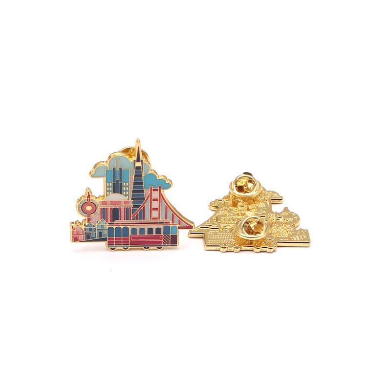 Op de afbeelding: Twee goudkleurige emaille pins met bezienswaardigheden van San Francisco. E&eacute;n pin toont de Golden Gate Bridge, een kabeltram en stadsgebouwen in rood, blauw en roze. De andere pin toont de achterkant met de pin sluitingen.