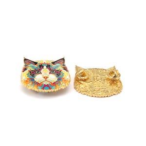 Puede incluir: Un pin de esmalte con forma de cara de gato colorido con un borde dorado. El pin presenta un dise&ntilde;o detallado con tonos de azul, naranja, amarillo y verde. La parte posterior del pin es dorada con dos cierres de alfiler.