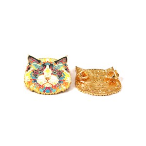 Puede incluir: Dos broches de esmalte con forma de gato. Un broche presenta un dise&ntilde;o colorido e intrincado con detalles amarillos, azules, rojos y negros. El otro broche es de color dorado con un dise&ntilde;o en relieve detallado. Ambos broches est&aacute;n sobre un fondo blanco.