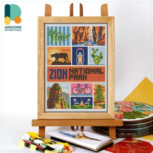 May include: A framed cross-stitch artwork featuring a collage of Zion National Park scenes. The design includes images of a mountain, a cougar, a lantern, cacti, a bicycle, and a campfire. The text "ZION NATIONAL PARK" is prominently displayed. The artwork is displayed on a wooden easel.