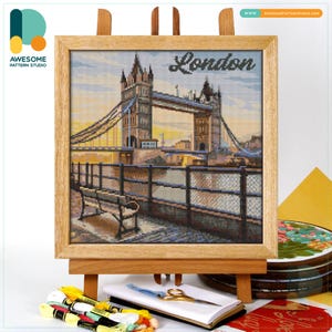 May include: Cross-stitch picture of Tower Bridge, London, in a light wood frame. The artwork is displayed on a wooden easel, with embroidery supplies including thread, scissors, and a notebook in the foreground. The word "London" is written at the top.