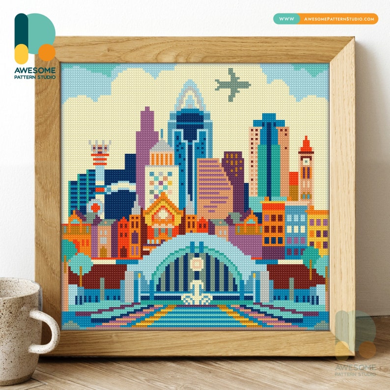 Op de afbeelding: Een kleurrijk pixel art kruissteekpatroon van een skyline met een groot gebouw in het midden en een klein vliegtuig dat erboven vliegt. Het patroon is ingelijst in een lichtbruin houten lijst.