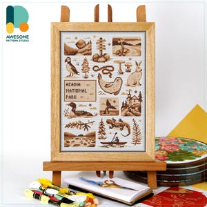 May include: A framed cross-stitch sampler featuring Acadia National Park icons, displayed on a wooden easel. The artwork includes images of wildlife, landmarks, and the park's name. Embroidery floss, scissors, and a notebook are in the foreground.
