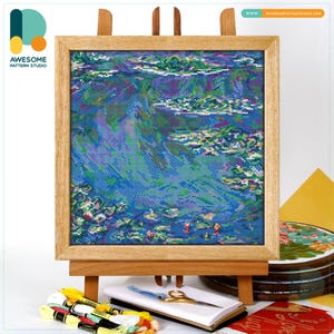 May include: A framed cross-stitch artwork depicting a water scene with blue, green, and purple hues. The artwork is displayed on a wooden easel. The frame is made of light-colored wood. The artwork is from Awesome Pattern Studio.