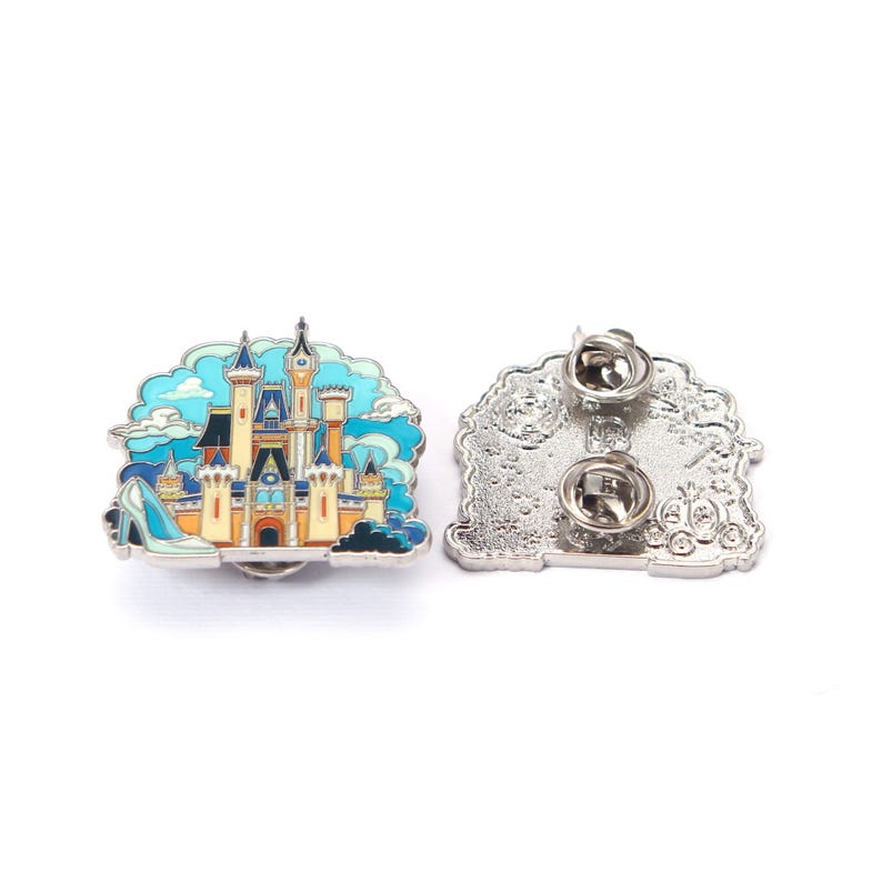 Pu&ograve; includere: Spilla smaltata con un design a castello colorato con sfondo cielo blu e una scarpetta di cristallo. La spilla ha un retro argentato con due perni.