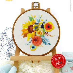 May include: Embroidered floral design in a wooden hoop, displayed on a small wooden easel. The embroidery features sunflowers, poppies, and other flowers in shades of yellow, orange, and blue. The text "Awesome Pattern Studio" is visible.
