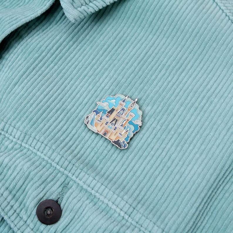 Pu&ograve; includere: Una spilla smaltata colorata con un castello da favola con dettagli blu, beige e bianchi. La spilla &egrave; applicata su una giacca di velluto a coste azzurra.