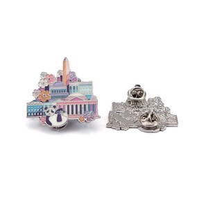 May include: A colorful enamel pin featuring a Washington D.C. cityscape with the Capitol Building, Washington Monument, and a panda bear. The pin is accented with floral details in shades of pink, purple, and orange. The back of the pin shows the pin's clasp.