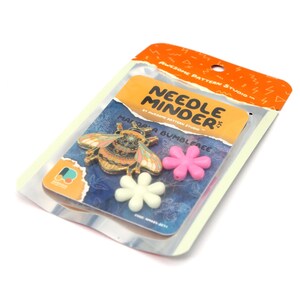 May include: A packaged needle minder set featuring a decorative bee pin and two flower-shaped magnets. The bee pin has intricate details with orange, pink, and gold accents. The packaging includes the text "Needle Minder" and "Mariposa Bumblebee".