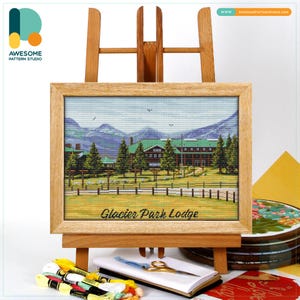Op de afbeelding: Een ingelijste kruissteeklandschap van Glacier Park Lodge, tentoongesteld op een houten ezel. Het kunstwerk toont een gedetailleerde weergave van de lodge, bomen en bergen. Het frame is van lichtgekleurd hout. Borduurgaren, schaar en een notitieboekje staan op de voorgrond.