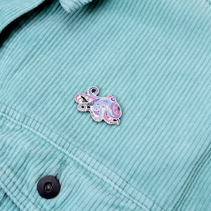 May include: A colorful octopus enamel pin with intricate patterns in shades of pink, purple, and blue. The pin is set against a light teal corduroy fabric background. The pin has a silver outline and a small loop at the top.