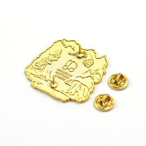 May include: A gold-colored pin with a detailed landscape design, including trees, mountains, and a bear. The pin has two pin backs and the text "AMERICAN EXPEDITION STUDIO".