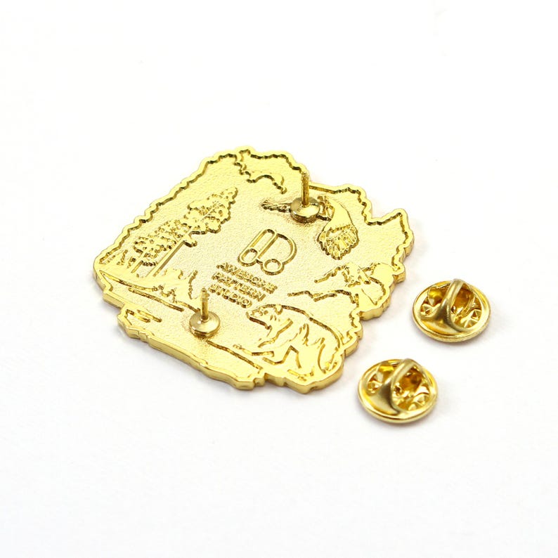 May include: A gold-colored pin with a detailed landscape design, including trees, mountains, and a bear. The pin has two pin backs and the text "AMERICAN EXPEDITION STUDIO".