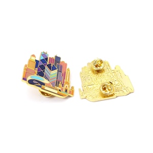 May include: A colorful enamel pin featuring a cityscape design with various buildings in shades of blue, purple, pink, and yellow. The pin has a gold-colored border and two pin backs. The design is a stylized representation of a city skyline.