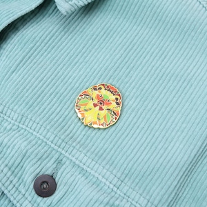 Puede incluir: Un pin decorativo de esmalte con forma de erizo con un borde dorado. El pin presenta un dise&ntilde;o detallado con tonos amarillos, naranjas, verdes y marrones. El pin est&aacute; colocado sobre una tela de pana de color verde azulado claro.