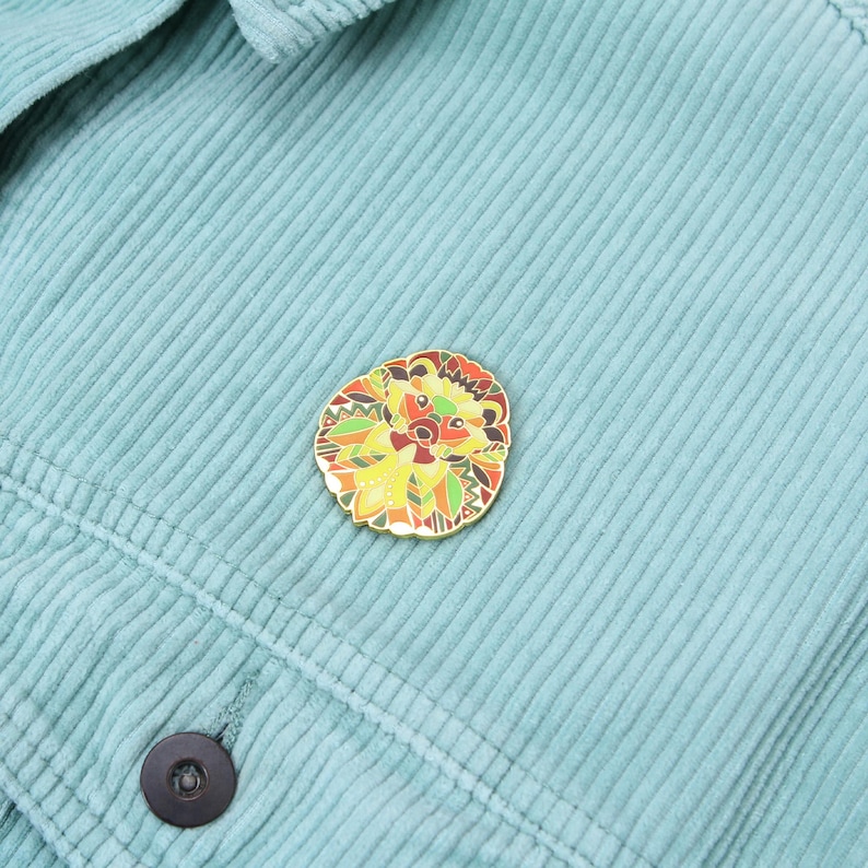 Puede incluir: Un pin decorativo de esmalte con forma de erizo con un borde dorado. El pin presenta un dise&ntilde;o detallado con tonos amarillos, naranjas, verdes y marrones. El pin est&aacute; colocado sobre una tela de pana de color verde azulado claro.
