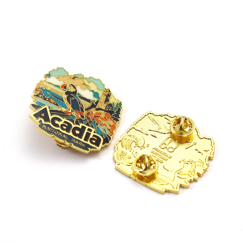 K&ouml;nnte beinhalten: Eine goldfarbene Emaille-Anstecknadel mit einer farbenfrohen Illustration des Acadia Nationalparks. Die Nadel zeigt einen Papageientaucher, einen Leuchtturm und den Text "Acadia National Park". Die R&uuml;ckseite der Nadel ist goldfarben mit zwei Nadelr&uuml;cken.