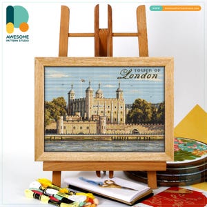 May include: A framed cross-stitch of the Tower of London, displayed on a wooden easel. The artwork features the historic landmark with a blue sky background. Embroidery floss, scissors, and a notebook are in the foreground.