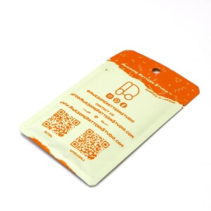 May include: A cream-colored package with an orange top and printed text. The package features the text "AWESOME PATTERN STUDIO" and social media icons. Two QR codes are visible, labeled "RETAIL" and "WHOLESALE".