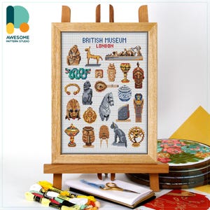 May include: Cross-stitch artwork of the British Museum in London, featuring various ancient artifacts. The framed piece is displayed on a wooden easel. The artwork includes a variety of colors and is surrounded by embroidery supplies, including thread and scissors.