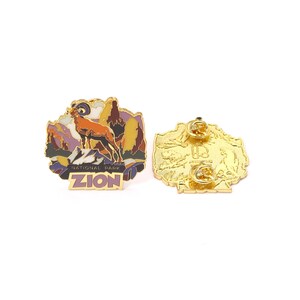 May include: A gold-colored enamel pin featuring a mountain ram in a landscape with the text "ZION NATIONAL PARK". The pin has a detailed design with shades of orange, purple, and yellow. The back of the pin is also gold-colored.