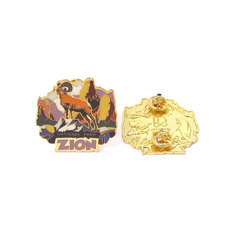 May include: A gold-colored enamel pin featuring a mountain ram in a landscape with the text "ZION NATIONAL PARK". The pin has a detailed design with shades of orange, purple, and yellow. The back of the pin is also gold-colored.