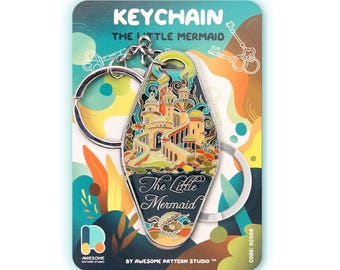 May include: A motel-style keyring featuring a colourful enamel illustration of an underwater castle and the text The Little Mermaid. The keyring is attached to a metal ring and presented on a branded backing card with abstract shapes.
