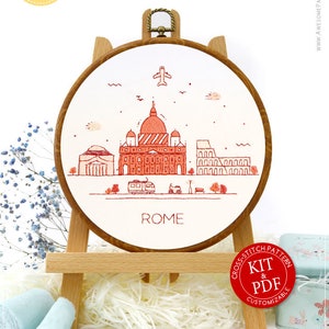 May include: A cross-stitch embroidery hoop featuring a red and white illustration of Rome's skyline, including the Colosseum and St. Peter's Basilica. The word "ROME" is below the image. The hoop is displayed on a wooden easel.