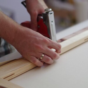 May include: A person using a red and silver staple gun to attach canvas to a wooden frame.
