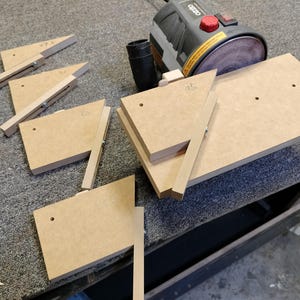 May include: A set of five wooden sanding blocks with different angles, designed for use with a sander. The blocks are made of light brown wood and have holes for attaching to a sander.