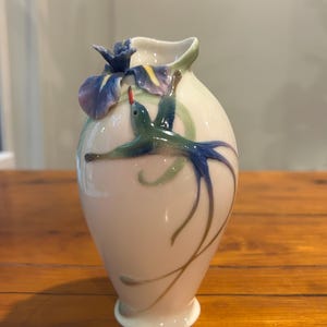 May include: A white ceramic vase featuring a hummingbird and iris design. The hummingbird is teal and blue with a red beak. The iris is purple and yellow. The vase has a curved shape and is on a wooden surface.