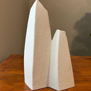 May include: A white, textured, abstract sculpture with two peaks. The sculpture has a rough, stucco-like surface and sits on a wooden table. The peaks are of varying heights, creating a modern, minimalist aesthetic.