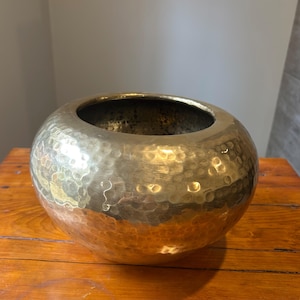 May include: A hammered brass bowl, spherical in shape with a wide opening. The surface is textured with small, irregular indentations. The brass has a warm, golden colour, typical of the material.