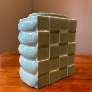 May include: A light green ceramic vase with a geometric design. The vase has a square base and a rectangular body with a series of raised squares and rounded ridges.