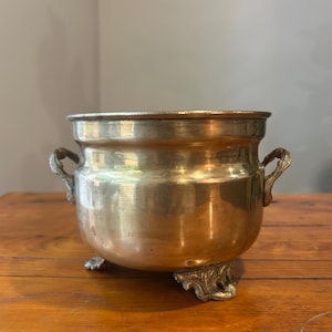 May include: A polished brass pot with a rounded body, featuring two ornate side handles and three decorative feet. The metal surface has a warm, golden metallic sheen with subtle reflections and a smooth, curved rim at the top opening.