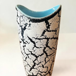 May include: A tall, tapered ceramic vase featuring a textured white crackle glaze over a black base layer. The vase has a distinctively curved, wave-like rim that reveals a smooth, bright light blue interior finish.