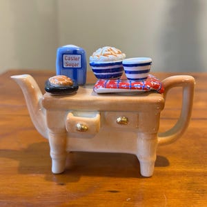 May include: A ceramic teapot shaped like a kitchen stove, with a tan body, white legs, and a curved handle. It features a blue and white container labeled "Caster Sugar", small bowls, and a plate. The teapot is on a wooden surface.