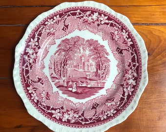 May include: A white ceramic plate with a pink floral design. The design features a central image of a building with trees and a fountain, surrounded by a border of leaves and vines.