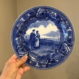 May include: A round ceramic plate featuring a blue and white transferware pattern. The center depicts a landscape with two figures standing near a body of water. The border is decorated with a detailed floral motif of roses and leaves.