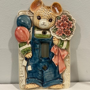 Peut inclure: Plaque de protection d'interrupteur en céramique représentant un ours en salopette bleue, tenant des ballons et des fleurs. L'ours a une fourrure brune et un visage blanc, sur un fond crème avec une bordure florale. Les ballons sont rouges et bleus.