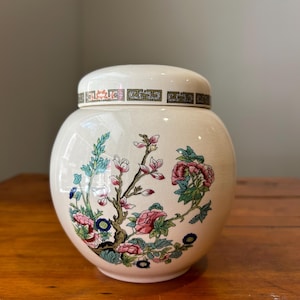 May include: A white ceramic jar with a floral design. The jar has a lid with a gold and black border. The flowers are pink, blue, and green.