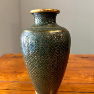 May include: A dark green vase featuring a repeating gold-coloured scale pattern across its surface. The vase has a narrow neck, a flared rim with a gold-coloured edge, and a smooth, rounded body that tapers towards the base, which also has a gold-coloured trim.