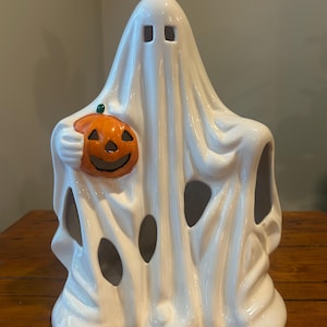May include: A white ceramic ghost figurine featuring a glossy finish, square eye cutouts, and a draped sheet appearance. The ghost holds a small, bright orange jack-o'-lantern with a carved face and a green stem in its right hand.