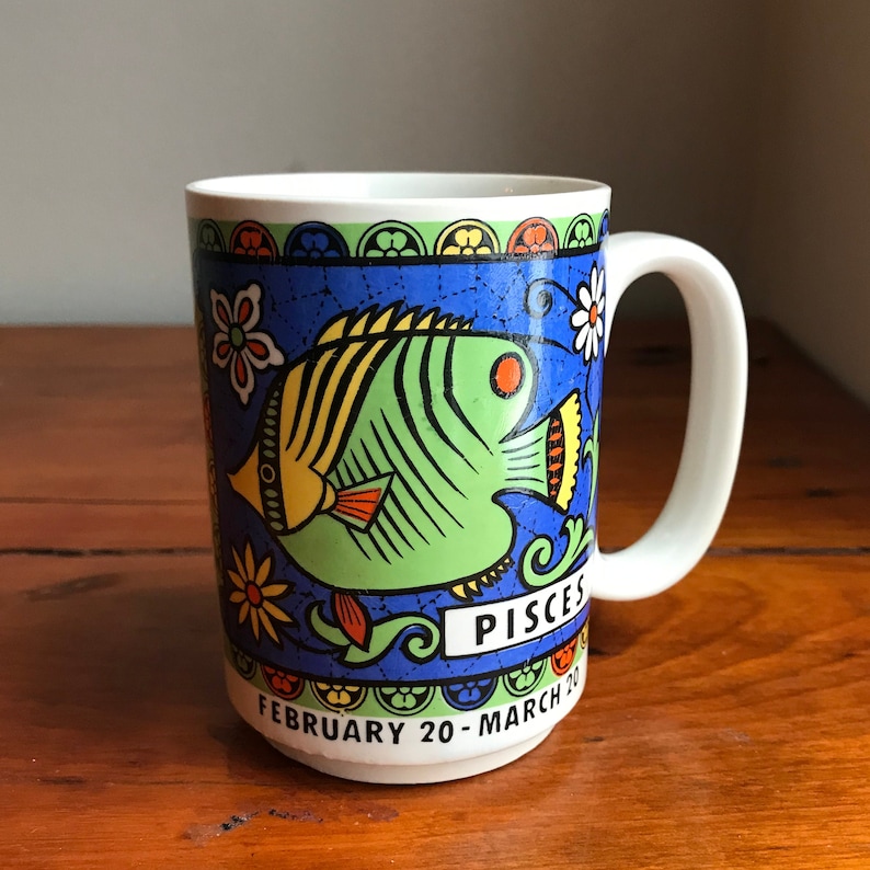 May include: A white ceramic mug with a blue, green, yellow, and orange design featuring a stylized fish and the words "Pisces" and "February 20 - March 20".