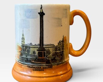 May include: A ceramic mug with an orange base and handle, featuring a black and white illustration of Trafalgar Square in London. The text 'TRAFALGAR SQUARE LONDON' is printed in black at the bottom of the illustration on the front of the mug.