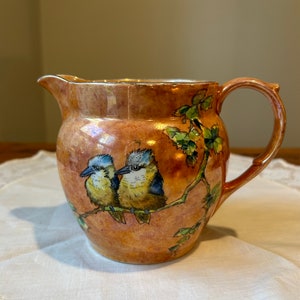 May include: A ceramic jug with a warm, mottled orange glaze. Two small birds with blue and yellow plumage perch on a branch with green leaves. The jug has a spout and a curved handle, ideal for serving.